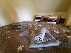 Een opgemaakt bed met een sprei met sterrenprint, twee kussens en een opgerolde handdoek op de vloer.
