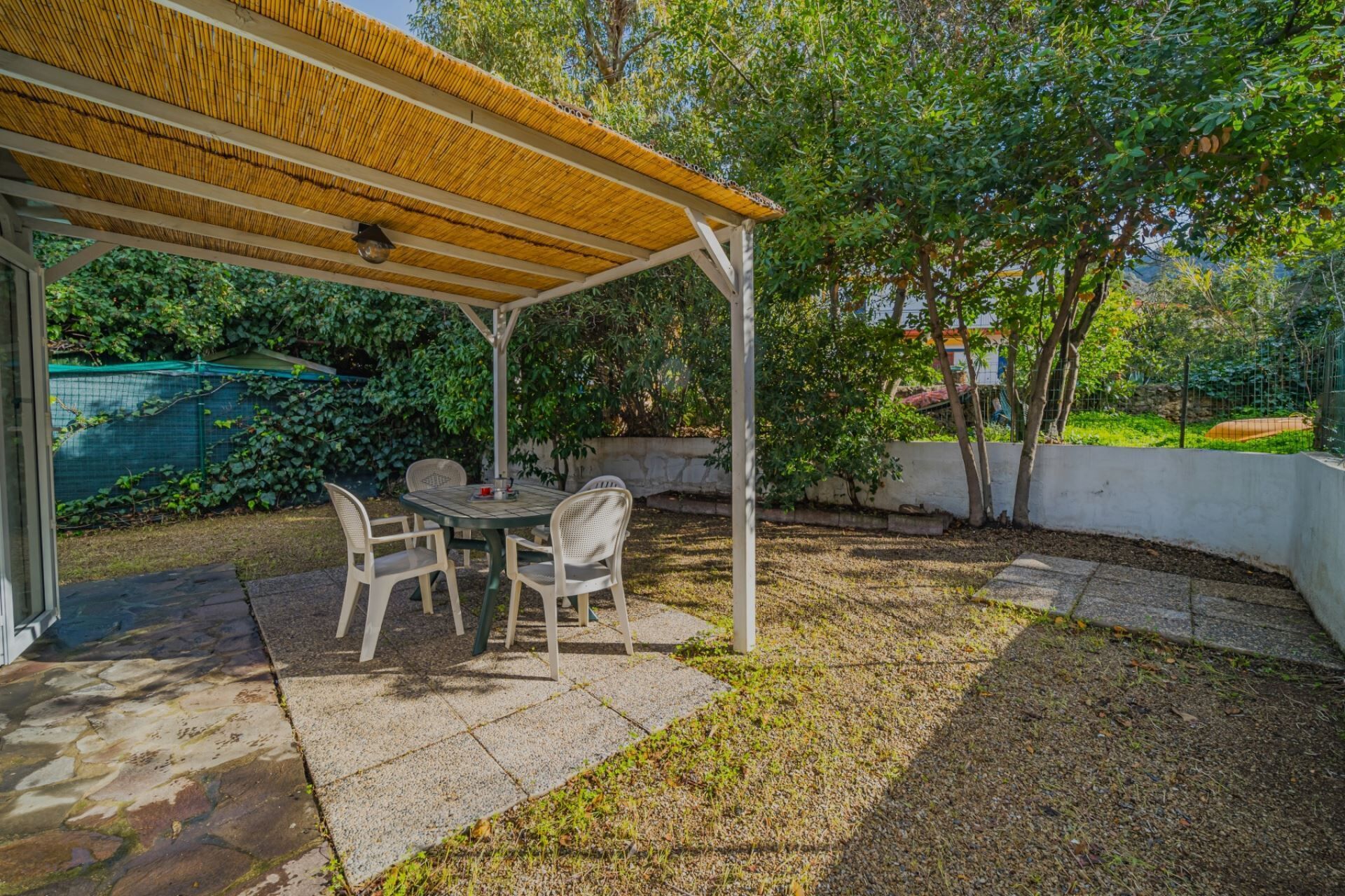 A covered patio with a table and chairs, surrounded by a garden and a white wall.