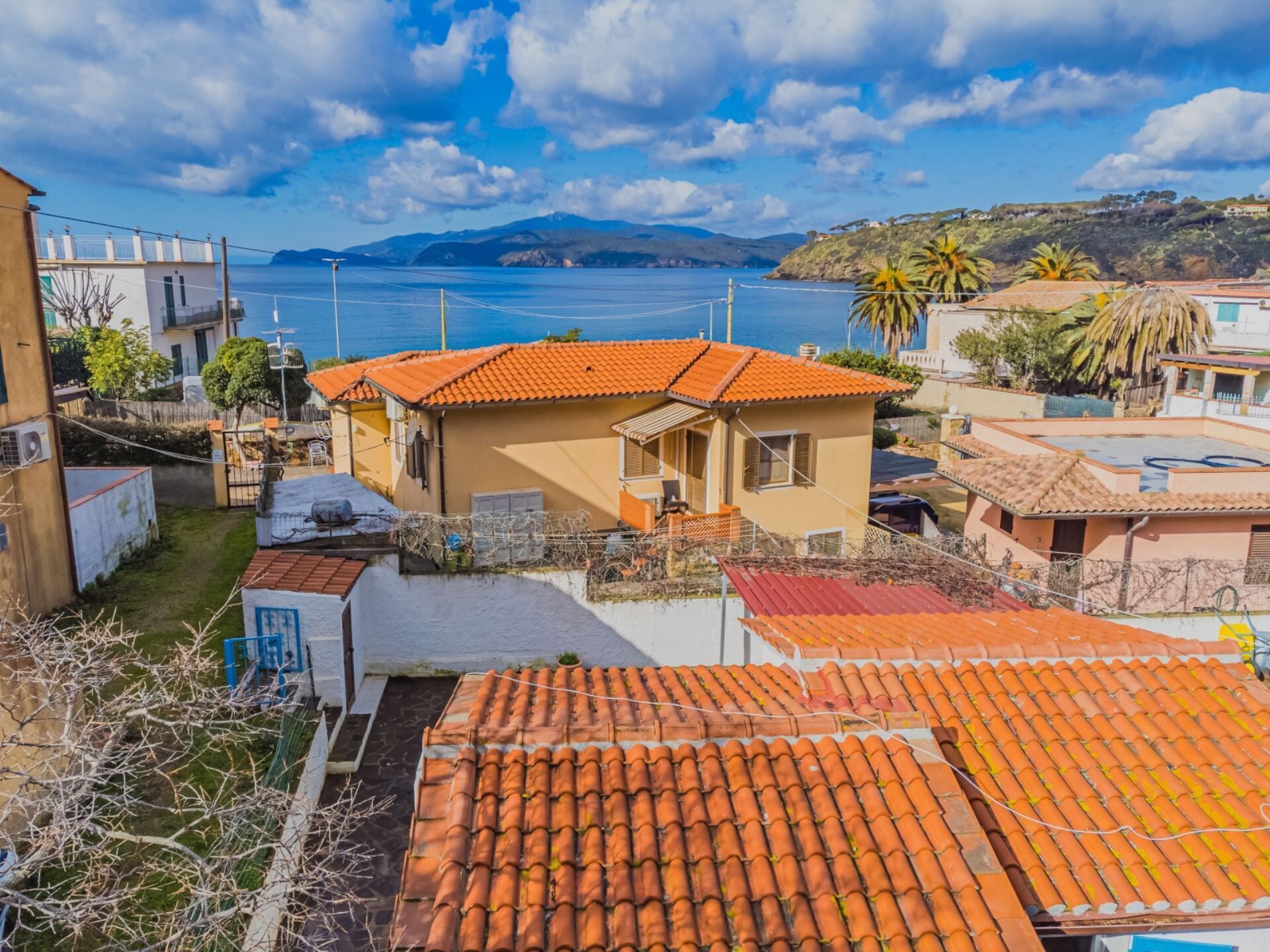 A coastal residential area with terracotta-roofed houses, a clear view of the sea, and a hilly landscape in the background.