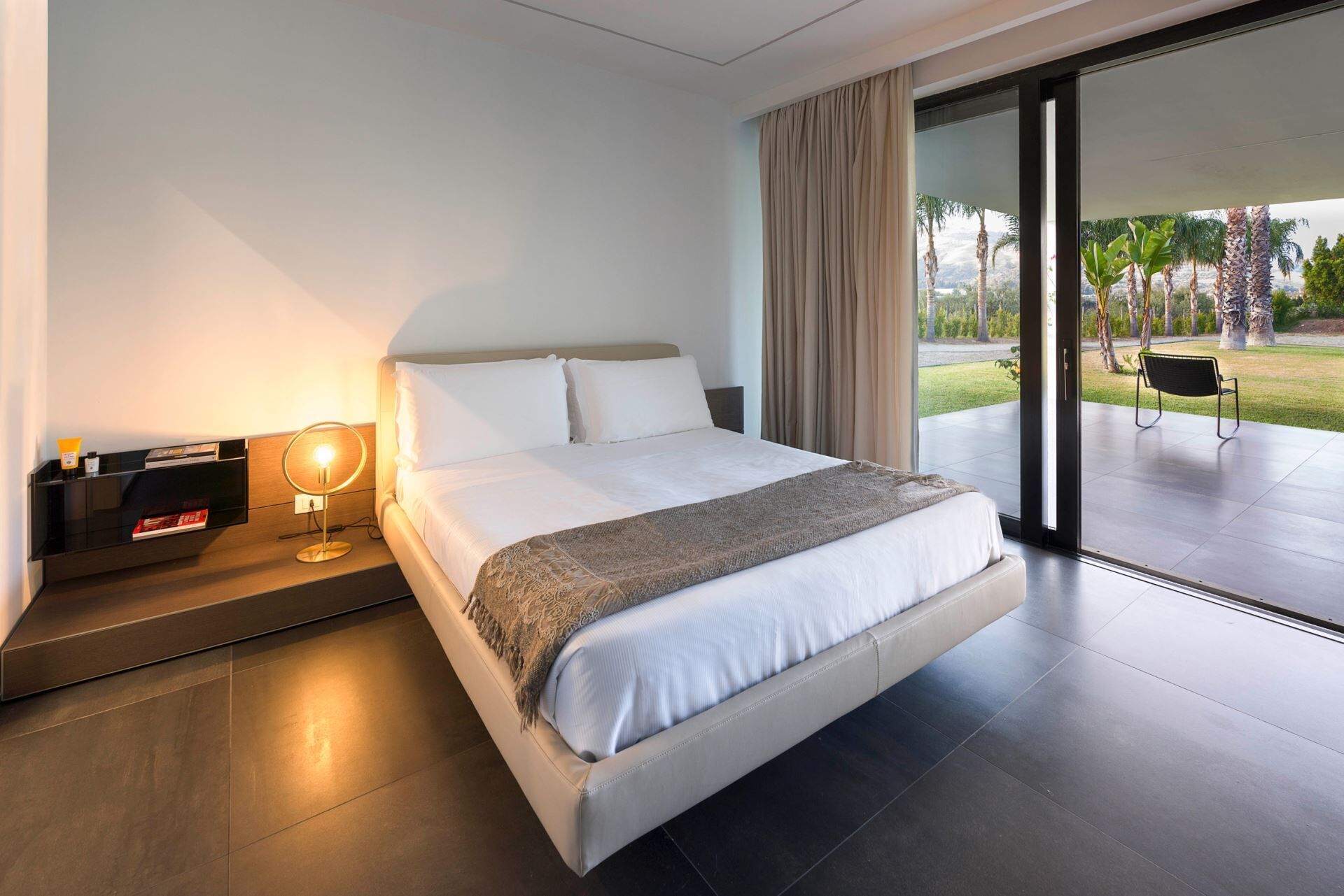 A modern hotel room with a large bed, a bedside table with a lamp, and a view of a garden through a sliding glass door.