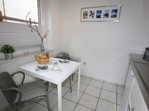 A small, clean kitchen with a white table set for two, a chair, a potted plant, and a framed picture on the wall.