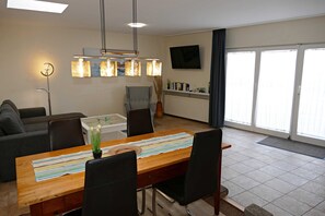 Een moderne woonkamer met een eettafel, stoelen, een bank, een televisie en een groot raam met zonwering.