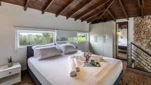A bedroom with a wooden ceiling, a bed with white linens, a nightstand, a window, and a stone wall. - EPTA Houses (KOS)