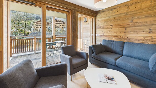 Un salon moderne avec un canapé, deux fauteuils et une table basse. De grandes fenêtres offrent une vue sur un balcon et les bâtiments environnants.