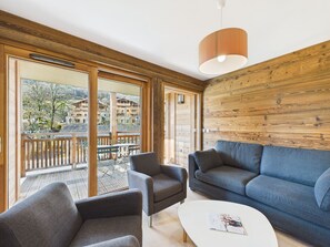 A modern living room with a sofa, two armchairs, and a coffee table. Large windows offer a view of a balcony and surrounding buildings.