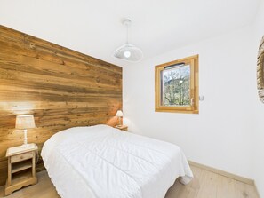 Un dormitorio con cabecera de madera, cama con edredón blanco, mesita de noche con lámpara y ventana con vistas a árboles.