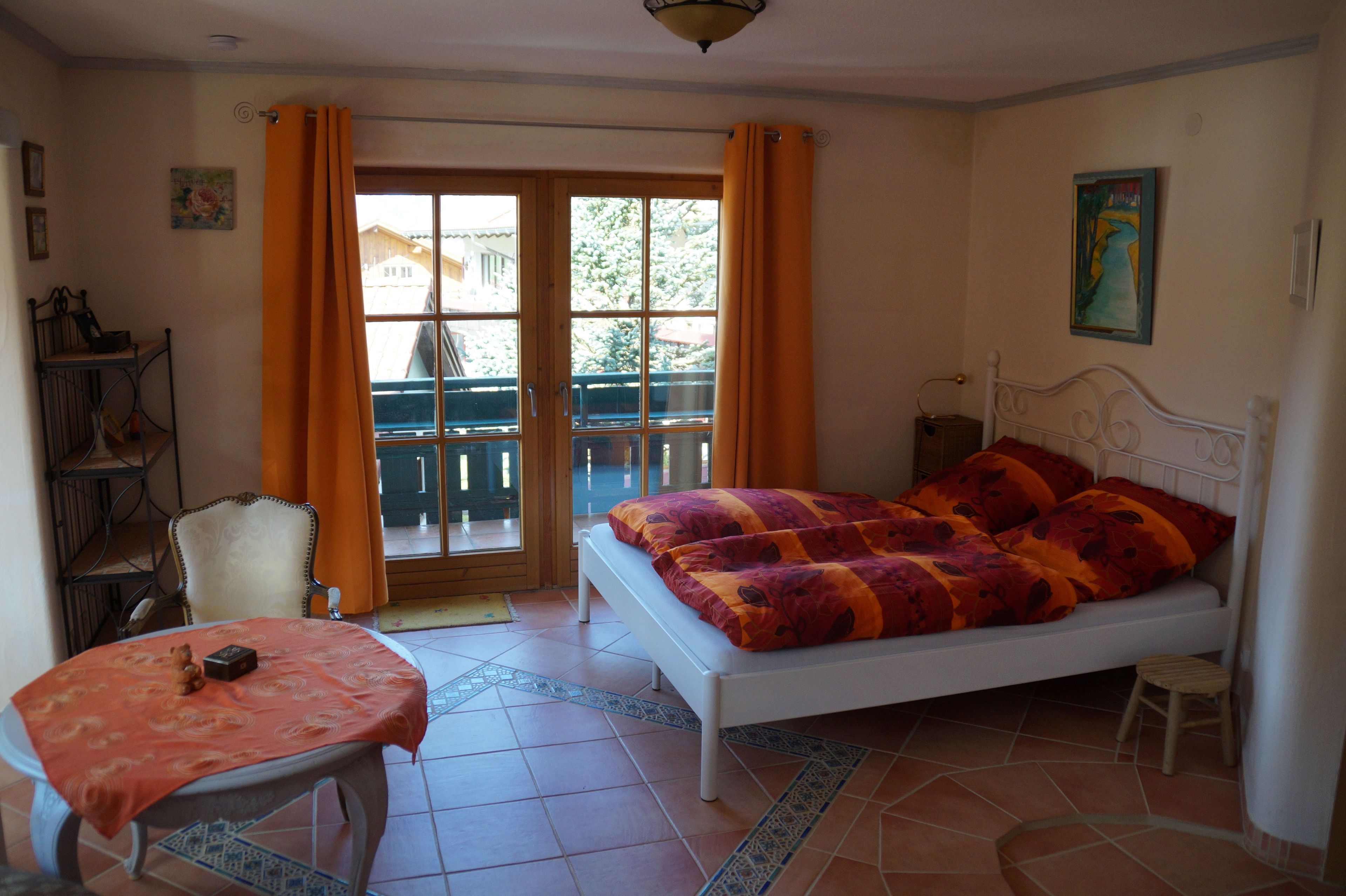 A bedroom with a bed, a table, a chair, a small stool, a bookshelf, and a window with curtains.