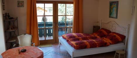 A bedroom with a bed, a table, a chair, a small stool, a bookshelf, and a window with curtains.