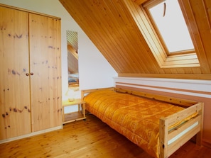 Una habitación pequeña de madera con techo inclinado y tragaluz. Tiene una cama, un armario y una mesita de noche con una lámpara.