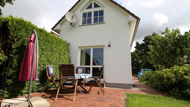 Een huis met twee verdiepingen, een schotelantenne, een terras met stoelen en een tafel, en een tuin.