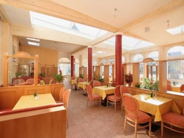 A restaurant with high ceilings, wooden beams, and large windows. Tables are set with yellow tablecloths and red chairs.