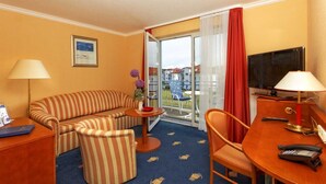 Ein Hotelzimmer mit einer gestreiften Couch, einem kleinen runden Tisch mit einer Vase, einem Sessel, einem Schreibtisch mit Lampe, einem Fernseher und einem Balkon mit rotem Vorhang.