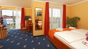 Ein Hotelzimmer mit Bett, Schreibtisch und Stuhl, einem kleinen Tisch mit Lampe, einem großen Fenster mit Vorhängen sowie einem Balkon mit Stuhl und Tisch.