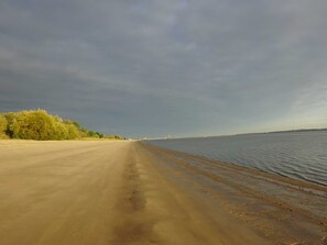 A beach with a road leading to the water, a distant bridge, and a cloudy sky.