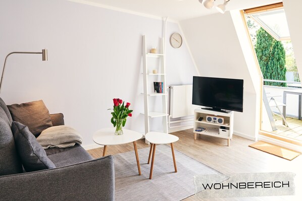 Un salon moderne avec un canapé, un téléviseur, une bibliothèque blanche et une petite table sur laquelle est posé un vase de fleurs.