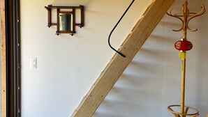 Una habitación con una escalera de madera apoyada en una pared, un espejo pequeño y una lámpara de pie con un adorno rojo.