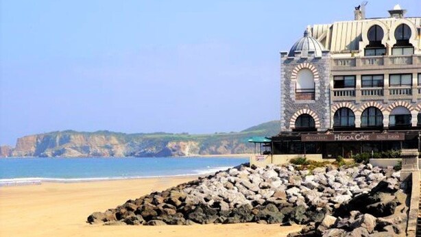 Ein Küstenhotel mit Steinfassade, Blick auf den Strand und einer felsigen Küstenlinie.