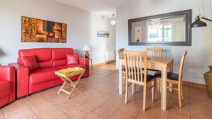 Ein Wohnzimmer mit rotem Sofa, einem Holzesstisch und Stühlen. Eine Lampe, ein kleiner Tisch mit einem Buch und Kunstwerken an den Wänden sind ebenfalls zu sehen.