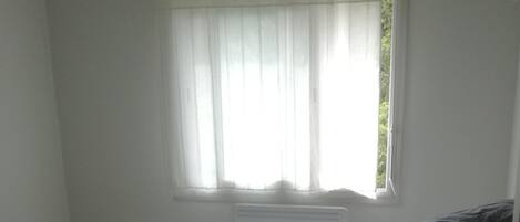 Ein Zimmer mit zwei Betten, einem Fenster mit durchsichtigen Vorhängen und einem Heizkörper über den Betten.