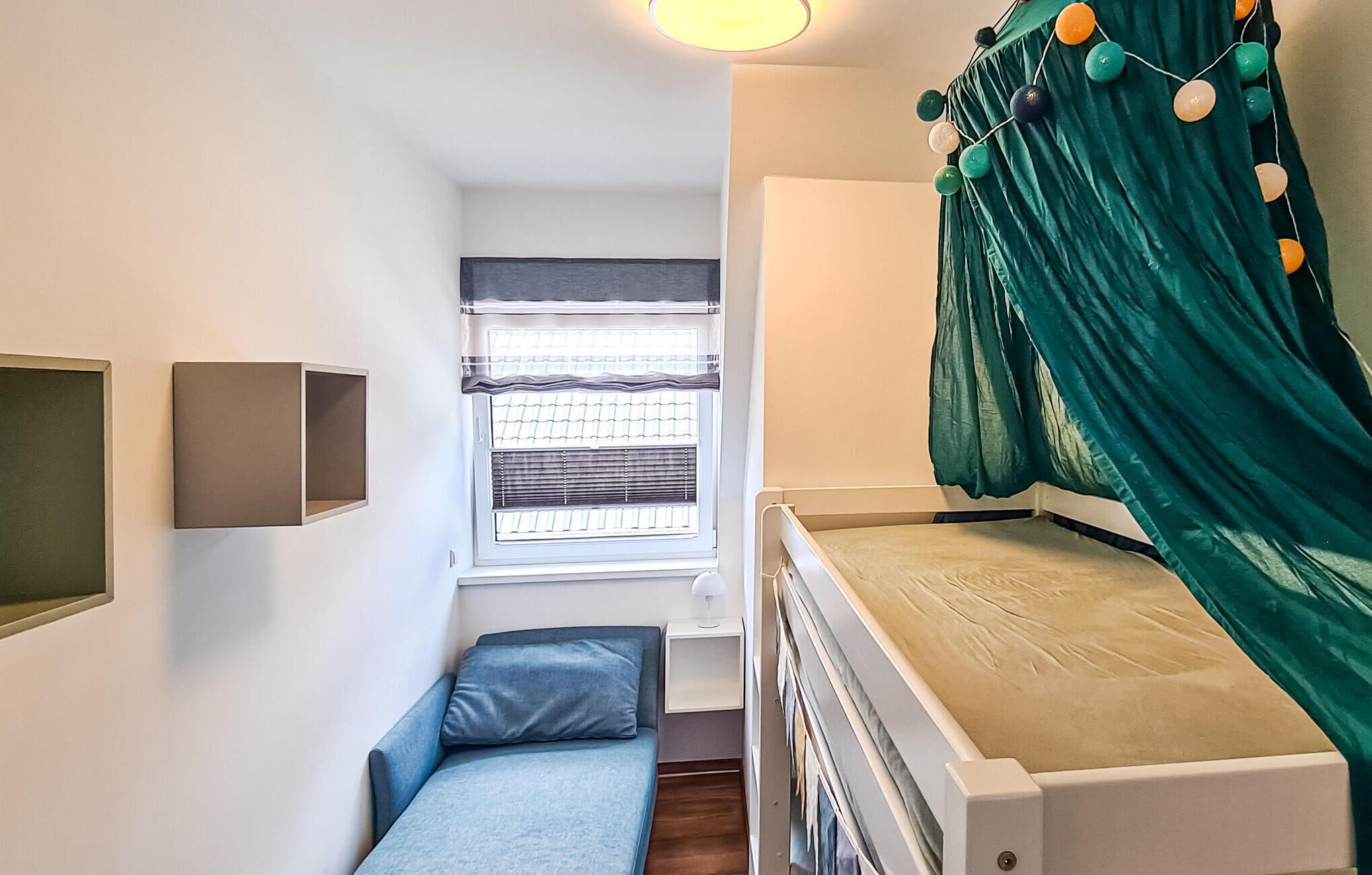 A compact room with a bed, a small seating area, a window with blinds, and a wall-mounted light fixture.