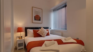 A neatly made bed with white and orange bedding, a red throw blanket, and two rolled white towels. There is a small bedside table with a lamp and a framed picture on the wall. - Apartment Sleeps 2 With Free Parking (Slough)
