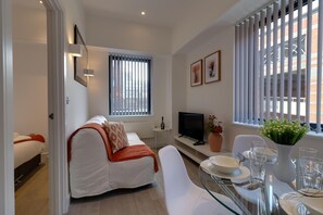 A modern living room with a sofa, a glass dining table set with plates and glasses, a TV, and a window with blinds. - Apartment Sleeps 2 With Free Parking (Slough)