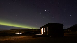 A small, dark building with a single illuminated window, set against a night sky with visible stars and a faint aurora. - Seljalandsfoss Cottage (Rangárþing eystra)