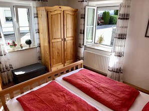 A bedroom with a bed, a wooden wardrobe, a bench, a window with curtains, and a view of the outside. - HoLiDay ApartMENT Allgäu (Ottobeuren)