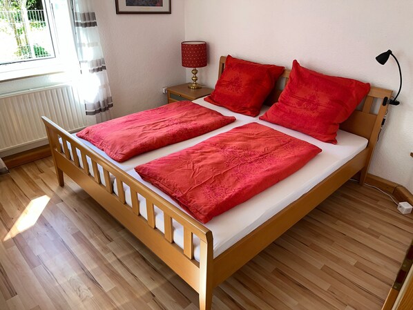 A wooden bed with red bedding in a room with wooden flooring and a window with curtains. - HoLiDay ApartMENT Allgäu (Ottobeuren)