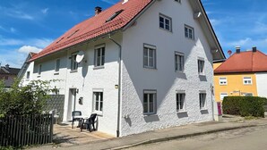 Een wit, twee verdiepingen hoog gebouw met een rood pannendak, meerdere ramen en een klein buitengebied met stoelen en een tafel.