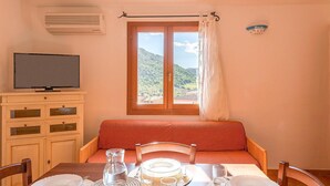 A cozy room with a bed, a dining table set for two, a television, and a window with a view of a mountainous landscape. - 1 bedroom nice apartment in Budoni (Budoni)