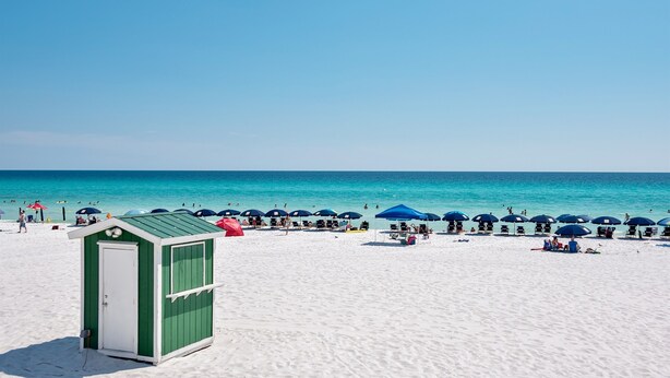 Una playa con una cabaña verde y blanca, sombrillas azules y arena blanca.