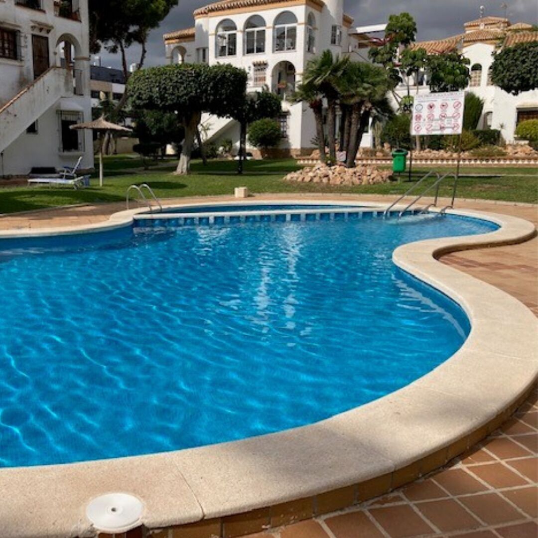 A swimming pool with a curved edge, surrounded by a paved area and a building with arched windows and a tiled roof.
