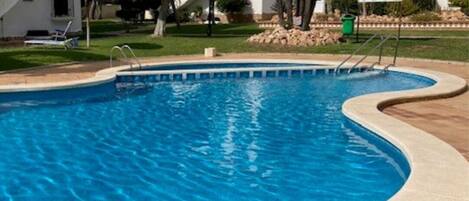 A swimming pool with a curved edge, surrounded by a paved area and a building with arched windows and a tiled roof.
