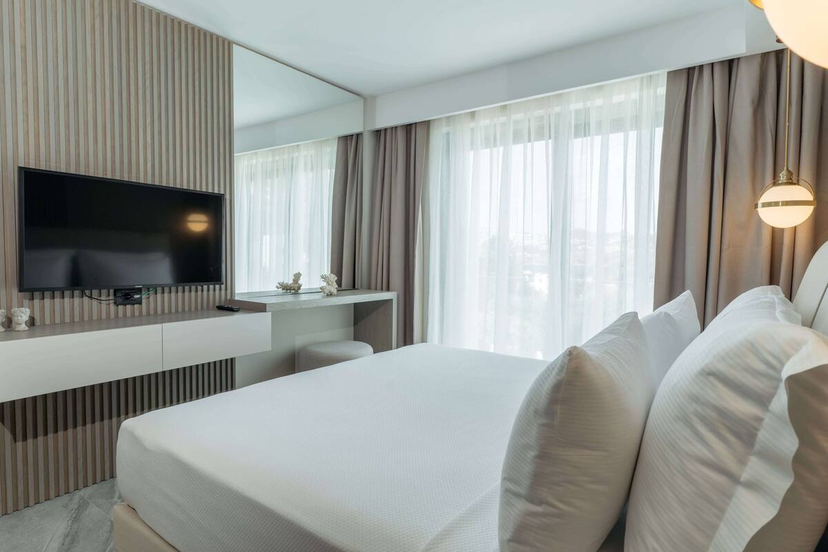 Una habitación de hotel moderna con una cama grande, un televisor de pantalla plana montado en un panel de madera, un escritorio pequeño con una silla y un ventanal con cortinas translúcidas.