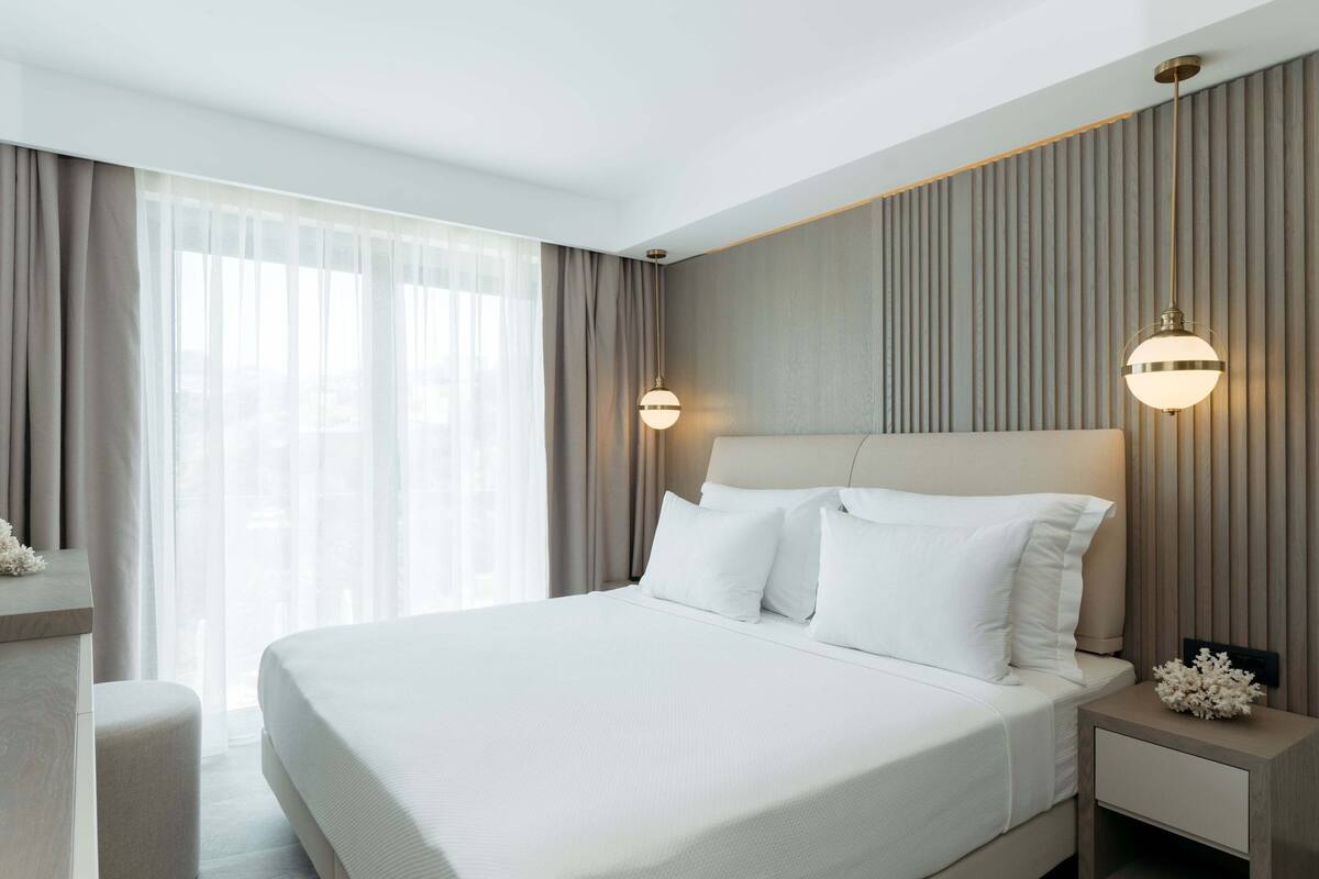 Una habitación de hotel moderna con una cama grande, una mesita de noche y un amplio ventanal con cortinas.
