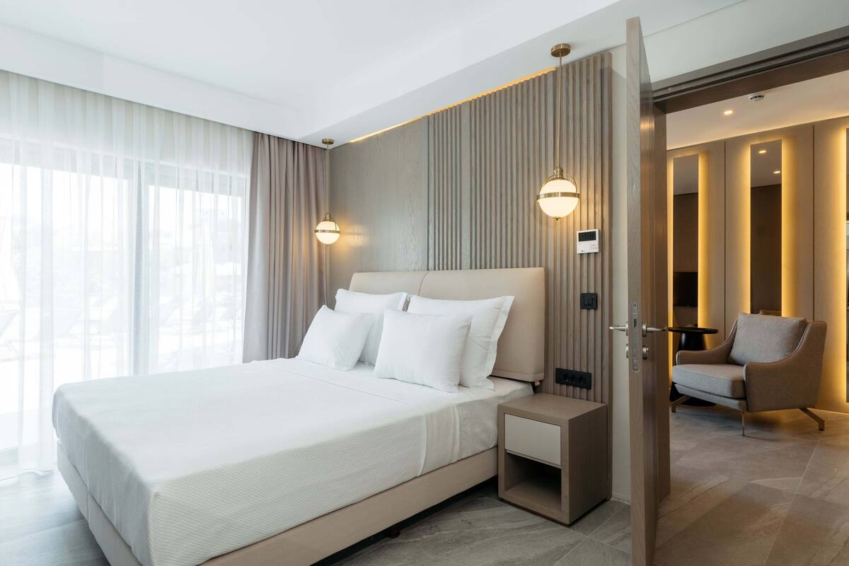 Habitación de hotel moderna con una cama grande, mesitas de noche, una silla y vistas a la ciudad.
