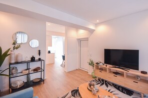 A modern living room with a large flat-screen TV, a wooden coffee table, and a blue sofa. There is a black shelving unit with decorative items and a potted plant.