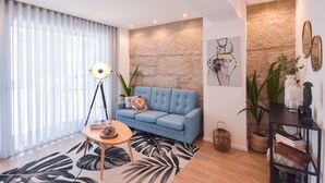 A modern living room with a blue sofa, a round coffee table, and a floor lamp. There are two large potted plants and a stone wall in the background.
