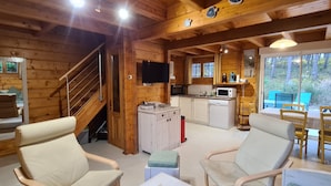 Interieur van een blokhut met een trap, een keukenhoek en een zithoek met stoelen en een tafel.