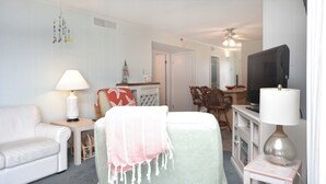 A living room with a white sofa, a small table with a lamp, a television, and a chair with a blanket.