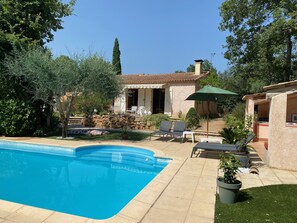Un espace piscine doté d’une terrasse en pierre, de chaises longues et d’un coin salon couvert avec un parasol.
