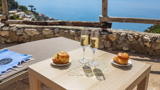 A table with two glasses of champagne and two muffins on a wooden table outdoors with a stone wall and a view of the sea in the background. - Villa Salice with private hot tub and solarium (Amalfi)