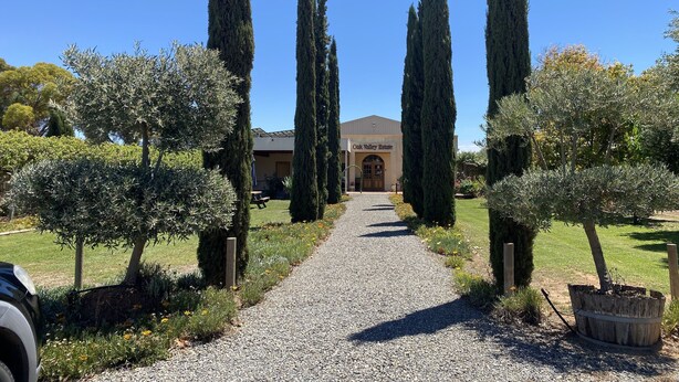 A gravel pathway leading to a building labeled "Oak Valley Estate" with tall cypress trees flanking the path.