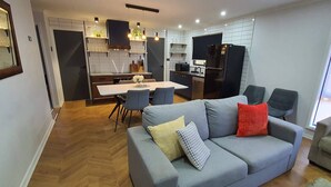 A modern living room with a grey sofa, a dining table, and a kitchen area with a black refrigerator.