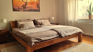 Un dormitorio con cama de madera, una mesita de noche, una lámpara, una ventana con cortinas y un cuadro colgado en la pared de un ciervo.