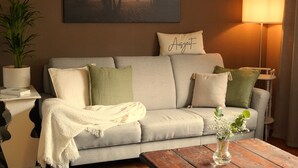 Una sala de estar con un sofá, almohadas, una manta, una mesa con un jarrón y un cuadro en la pared.