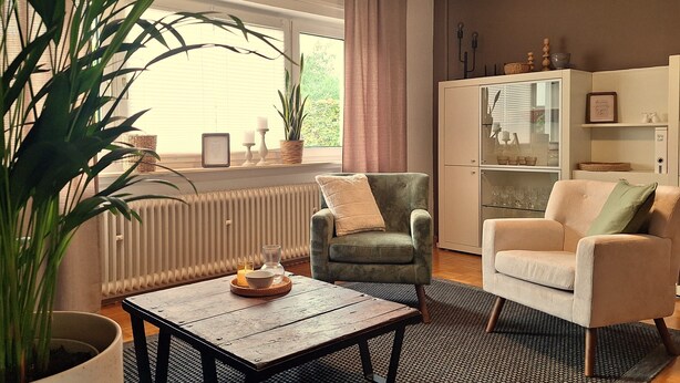 Una sala de estar con un sofá, un sillón y una mesita de centro. Hay una planta en maceta, un radiador y una ventana con cortinas.