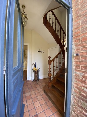 Un couloir avec une porte bleue, un sol carrelé, un escalier en bois et un luminaire fixé au mur.