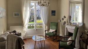 A room with a wooden floor, a green armchair, a wooden chair, a small table, a chandelier, and a large mirror.